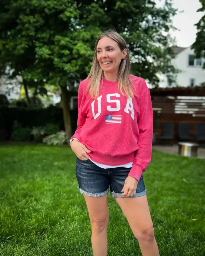 IMG_7927 - Sweet Motherly A woman standing in the grass wearing shorts and an american flag sweatshirt.
