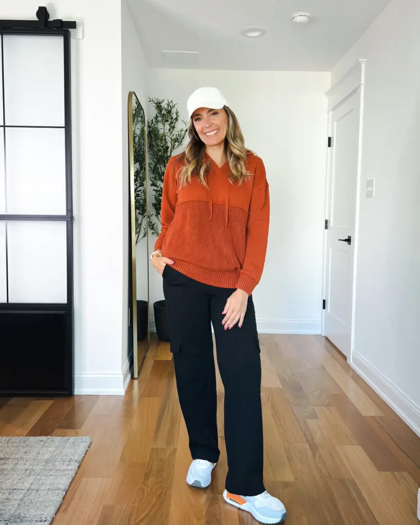 A woman standing in front of a door wearing an orange sweater.