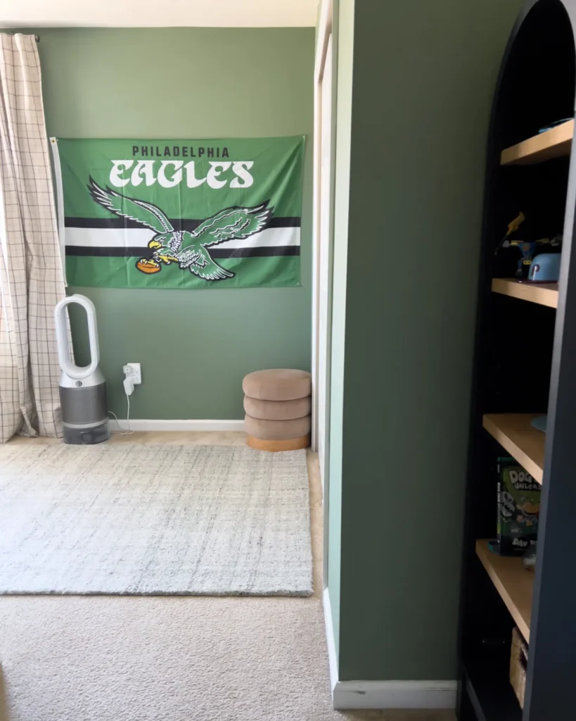Philadelphia Eagles flag on green bedroom wall with neutral plaid curtains, ivory rug, Dyson fan, and tan ottomans in a sports-themed boys’ room.