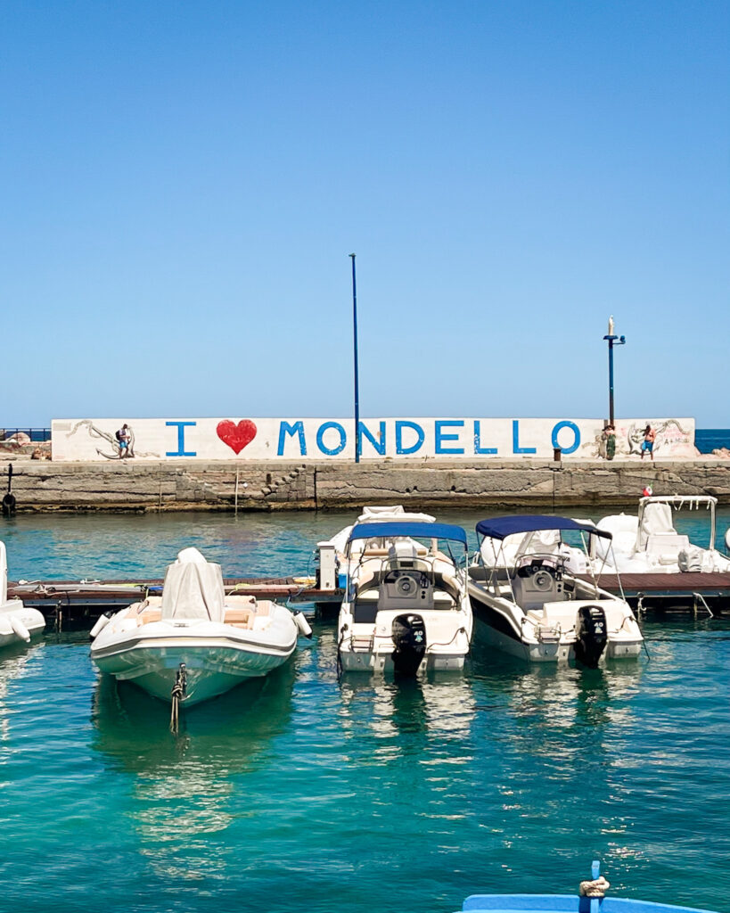Boats Docked in Mondello Harbor - Sweet Motherly Boats docked in Mondello’s turquoise harbor in front of an “I love Mondello” mural under a clear blue sky.
