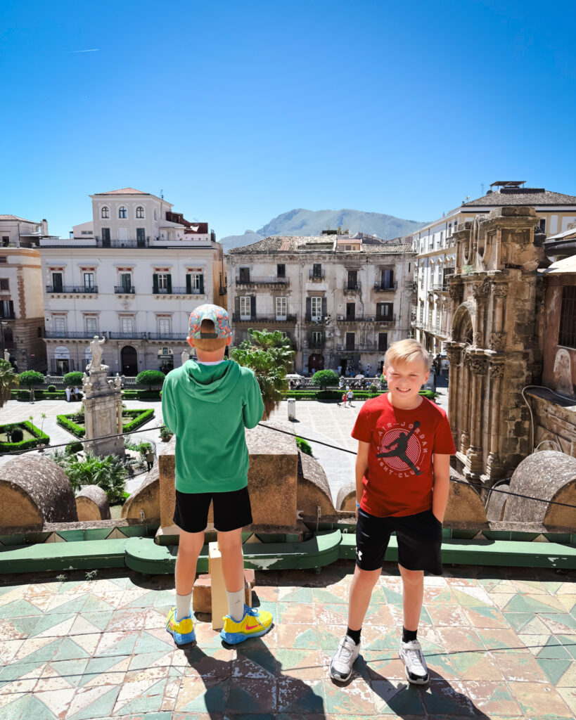 Palermo City Overlook with Kids at Scenic Piazza - Sweet Motherly Two boys overlooking a scenic piazza in Palermo with historic buildings, palm trees, and mountains in the distance.