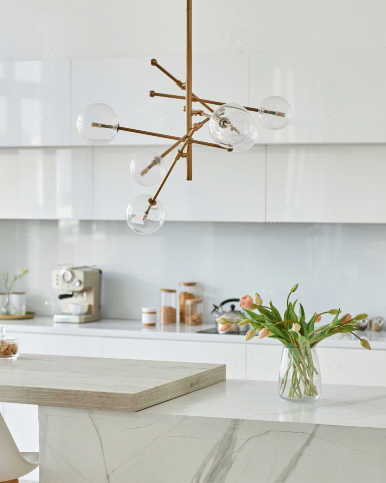 Bright modern kitchen with white cabinets, a marble waterfall island, a gold and glass globe chandelier, a glass vase of tulips, and a coffee station on the countertop.