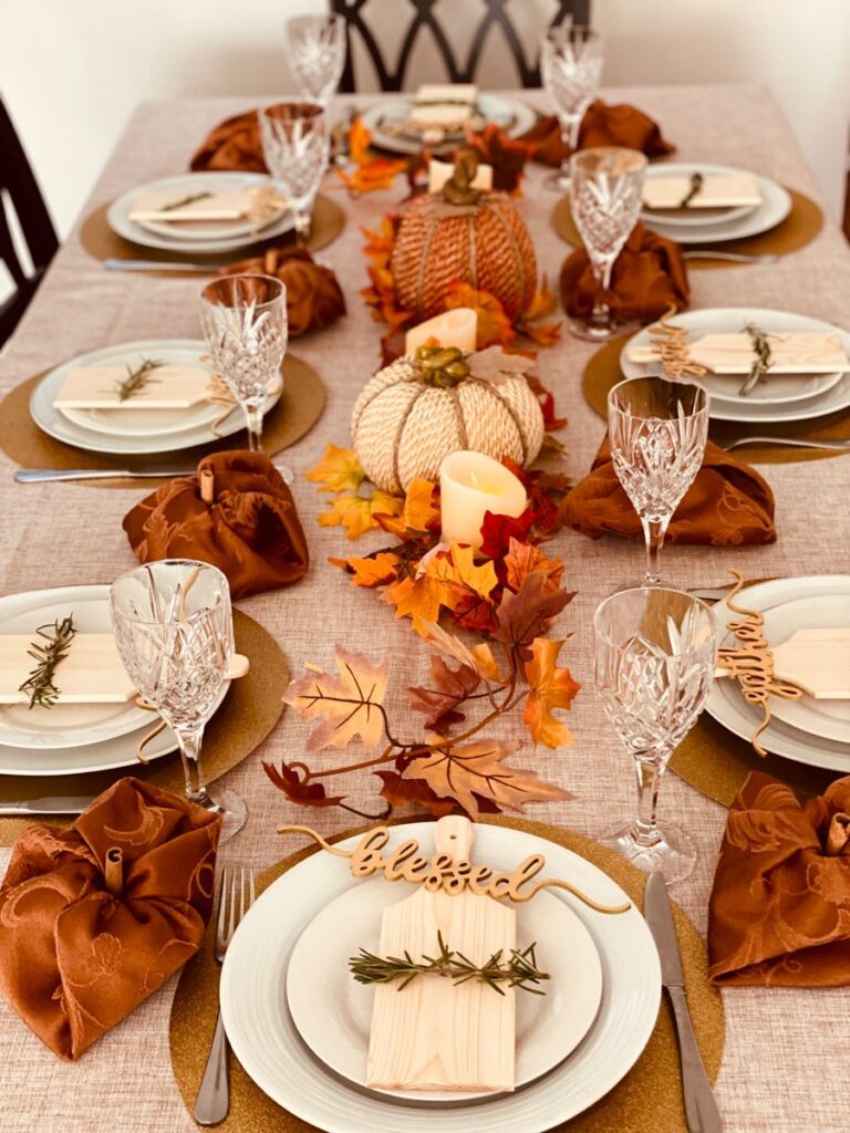 A fall-themed dining table set with crystal glasses, faux pumpkins, leaf garlands, and scripted wooden place settings on a neutral linen tablecloth.