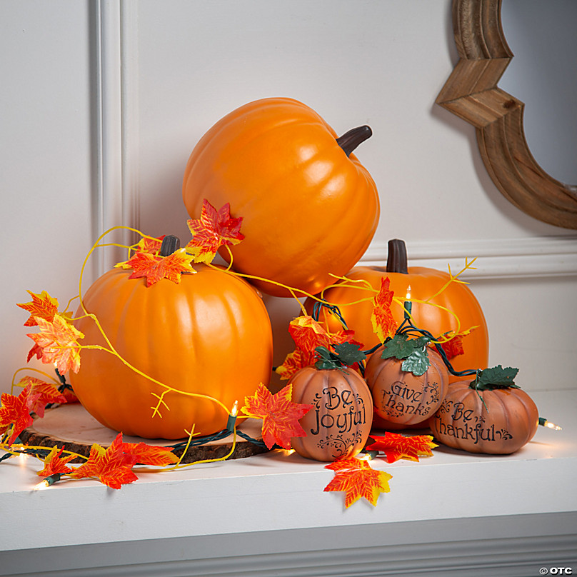 A display of bright orange faux pumpkins decorated with artificial fall leaves and string lights on a white mantel.