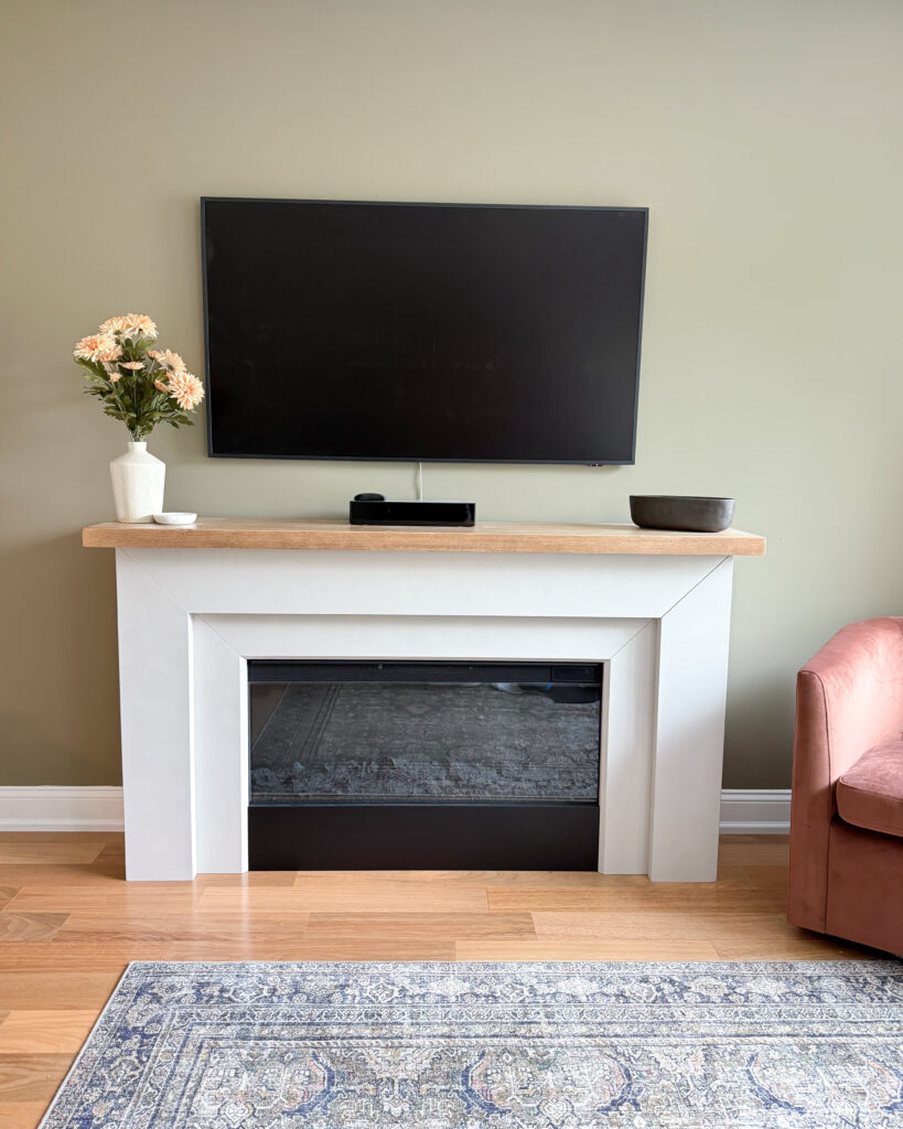 IMG_1316 - Sweet Motherly White fireplace with TV, wood mantel, blush florals, and neutral decor.