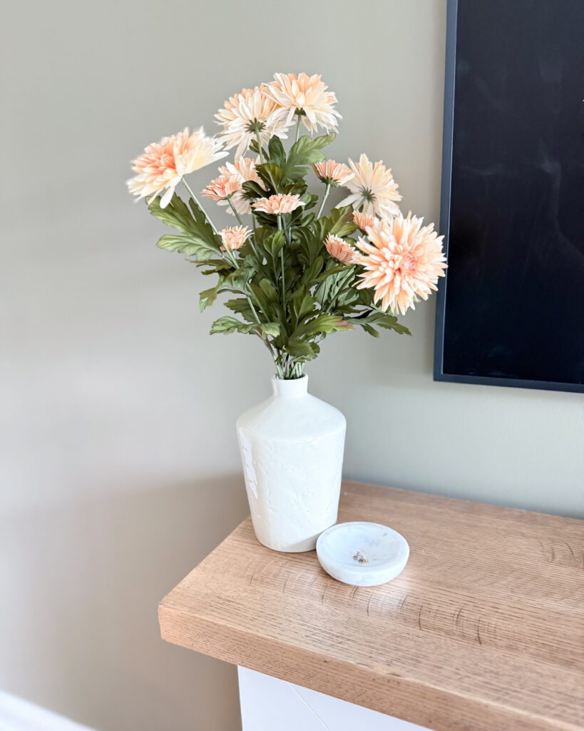 Close-up of blush faux daisies in a white vase on wood mantel.