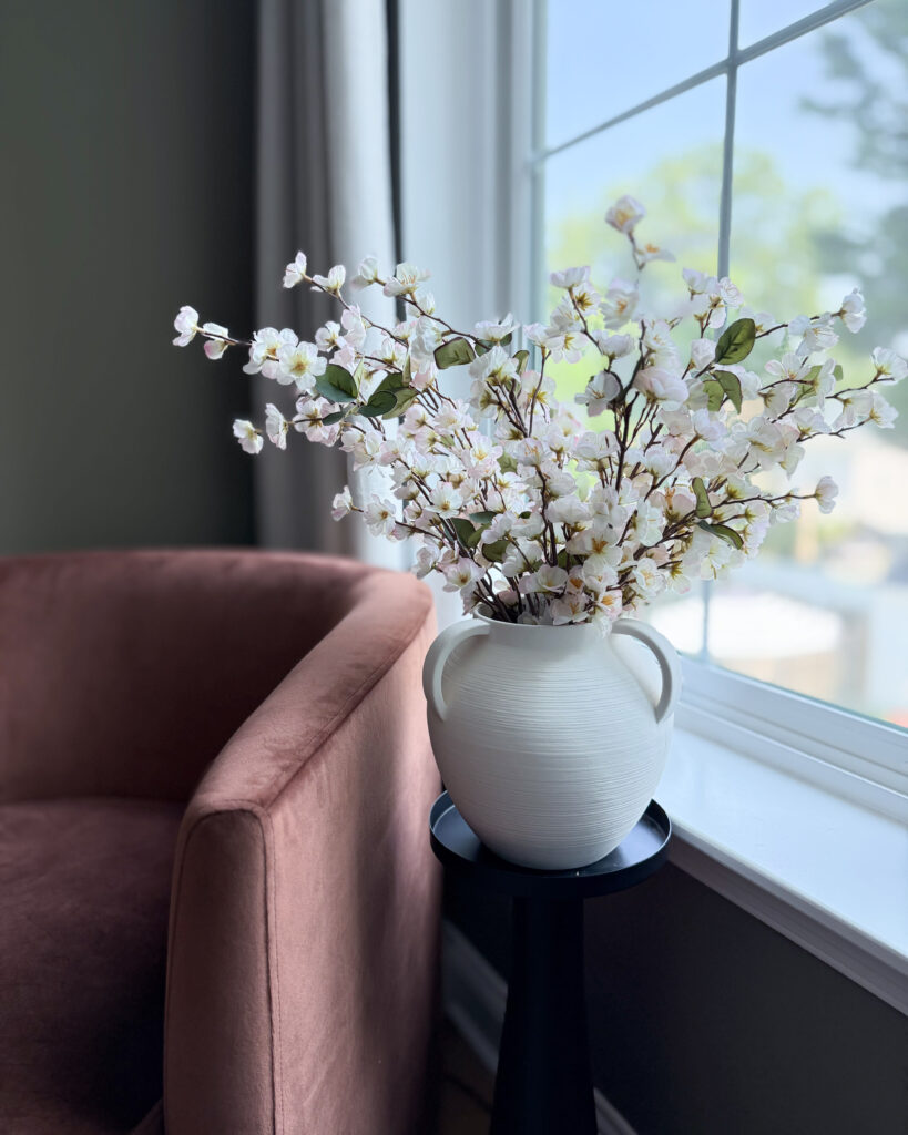 White vase of cherry blossoms on a black pedestal next to a blush chair by the window.