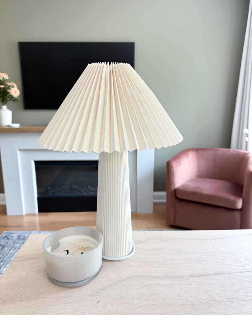 Pleated cream lamp and white candle on a wood mantel near a blush chair.