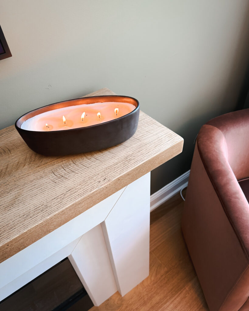 Five-wick oval candle on a light wood mantel next to a velvet pink chair.