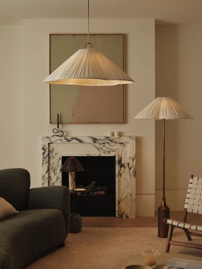 A warm-toned living room featuring pleated pendant and floor lamps, a marble fireplace, and cozy textured seating.