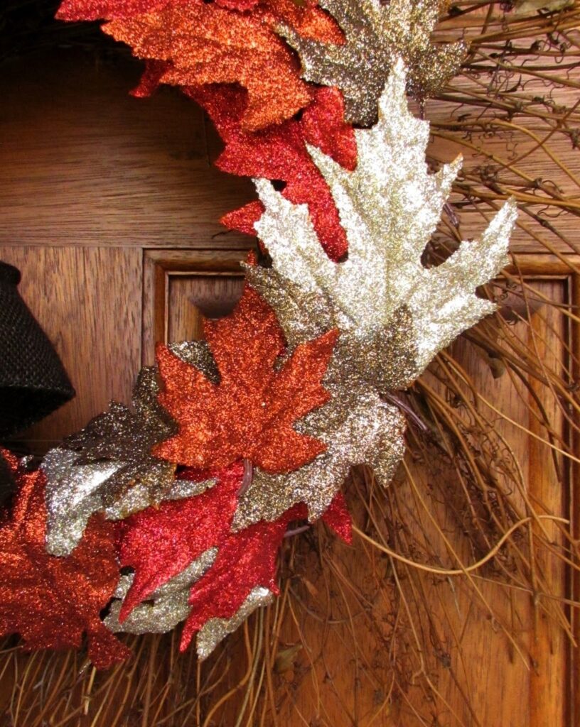 A close-up of a fall wreath decorated with glitter-covered red, gold, and bronze faux leaves on a wooden door.