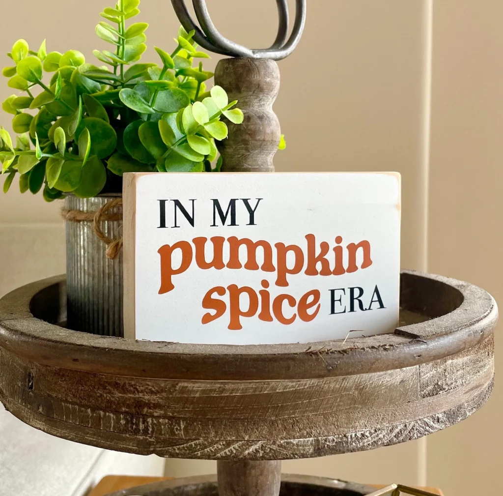 A small decorative wooden sign that reads “In My Pumpkin Spice Era” placed on a rustic tiered tray with a potted plant.