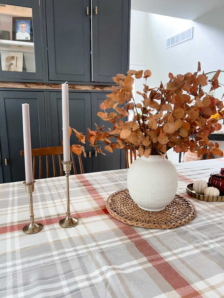 Fall-inspired dining table styled with a plaid tablecloth, brass candlesticks, a white vase with dried leaves, and small decorative pumpkins.
