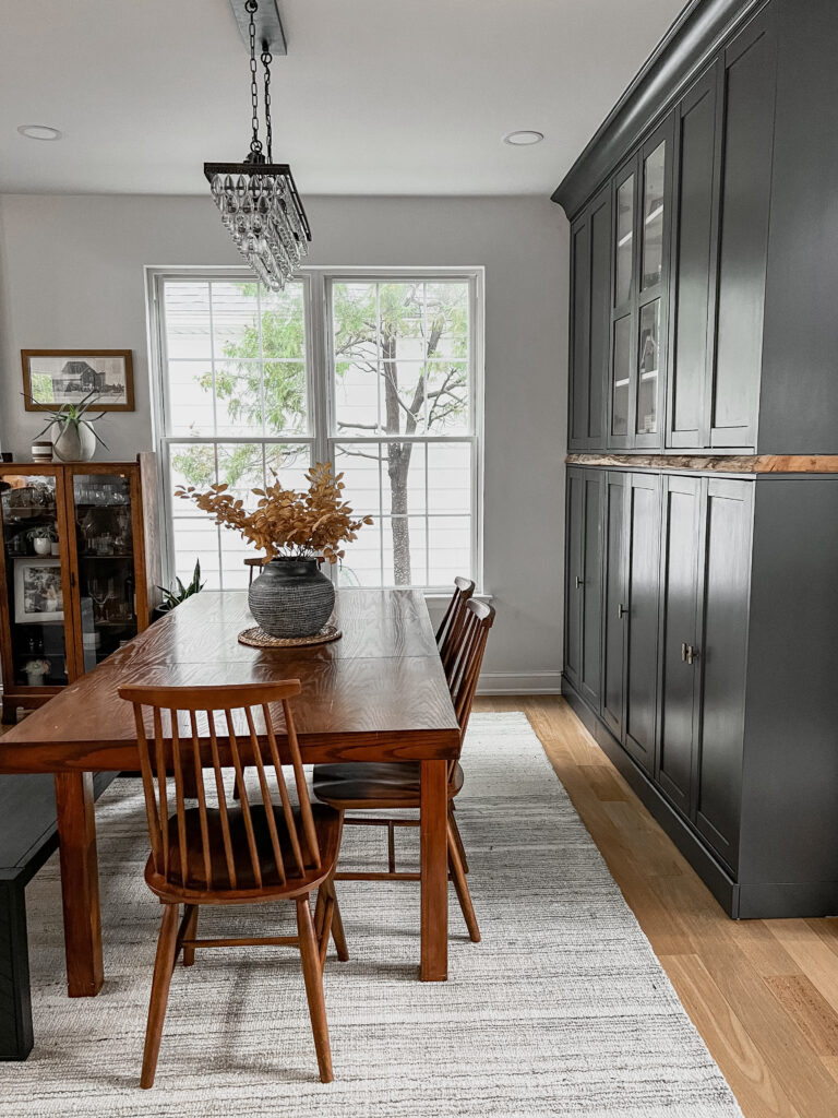 Cozy dining room featuring a wooden table with a dark ceramic vase and autumn branches, set against tall charcoal built-in cabinets and large windows.