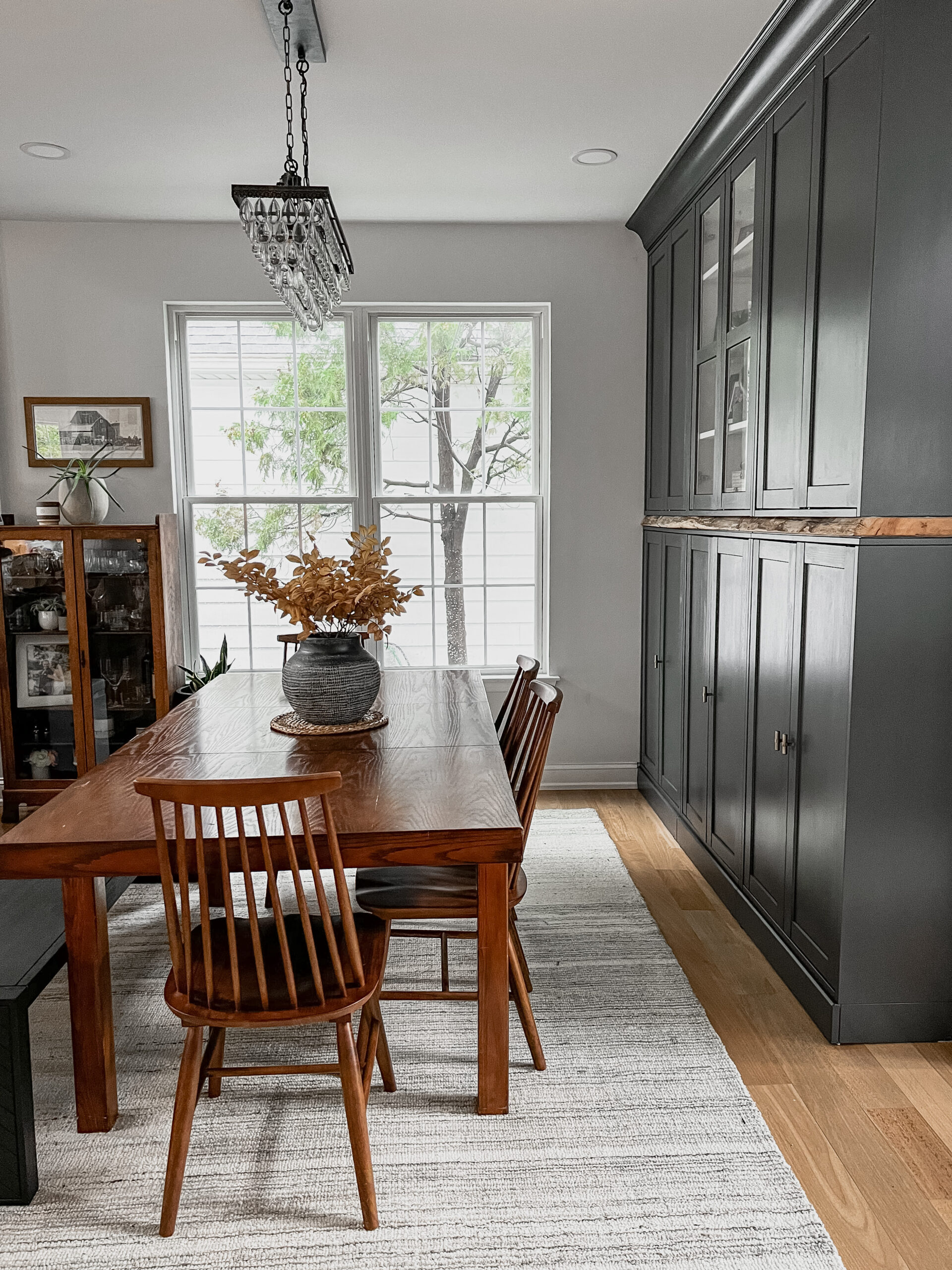 Cozy dining room featuring a wooden table with a dark ceramic vase and autumn branches, set against tall charcoal built-in cabinets and large windows.