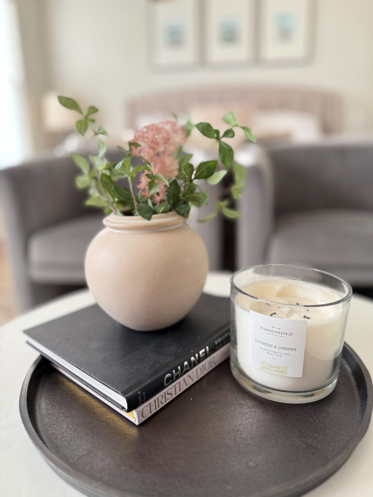 A close-up of a round tray styled with a beige vase of flowers, a white candle, and a stack of coffee table books for a cozy, layered touch.