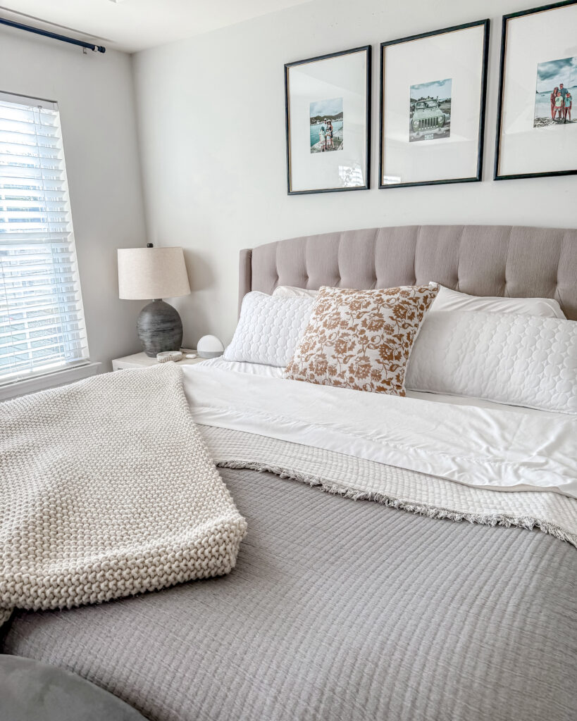 A cozy, layered bed featuring soft white sheets, textured knit blankets, and decorative pillows in neutral tones beside a gray nightstand and lamp.