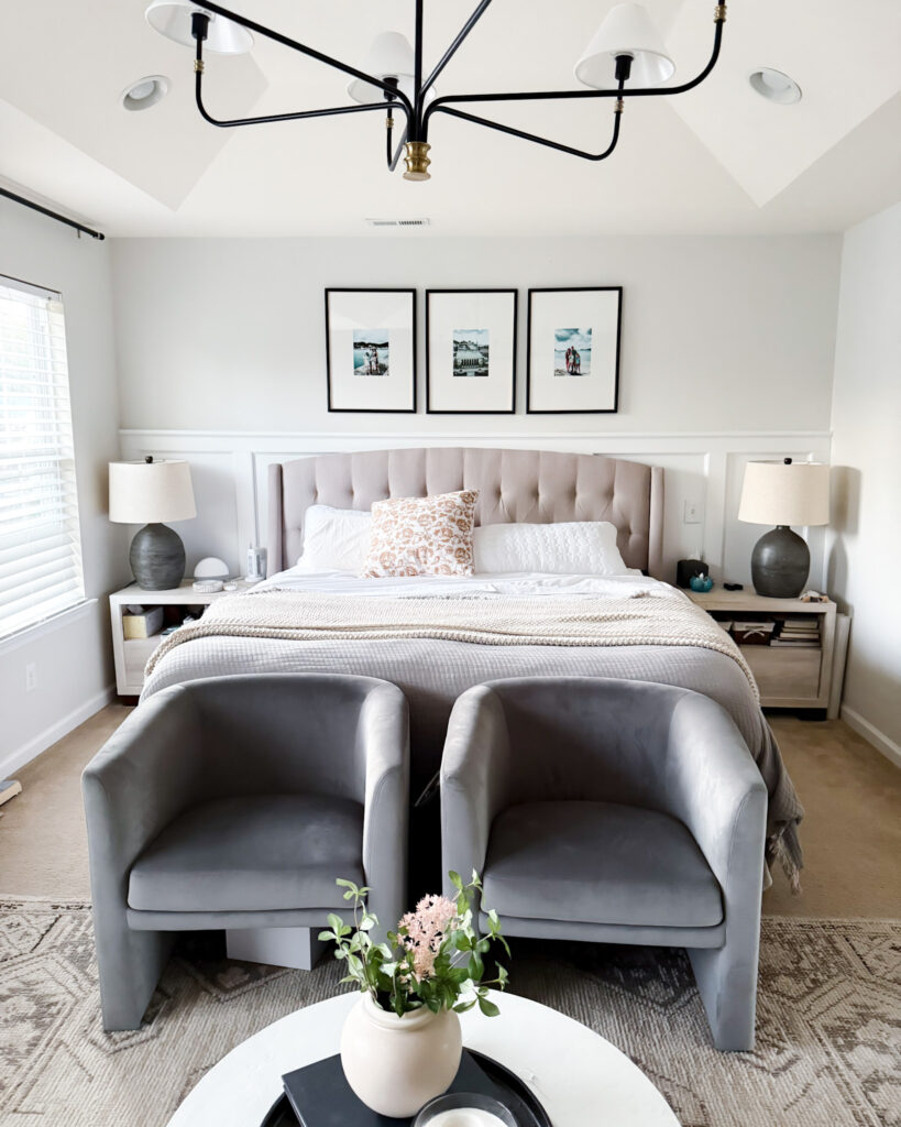 A modern, airy bedroom with soft gray chairs, a tufted bed, layered white bedding, neutral decor, and framed art above the headboard. Master Bedroom