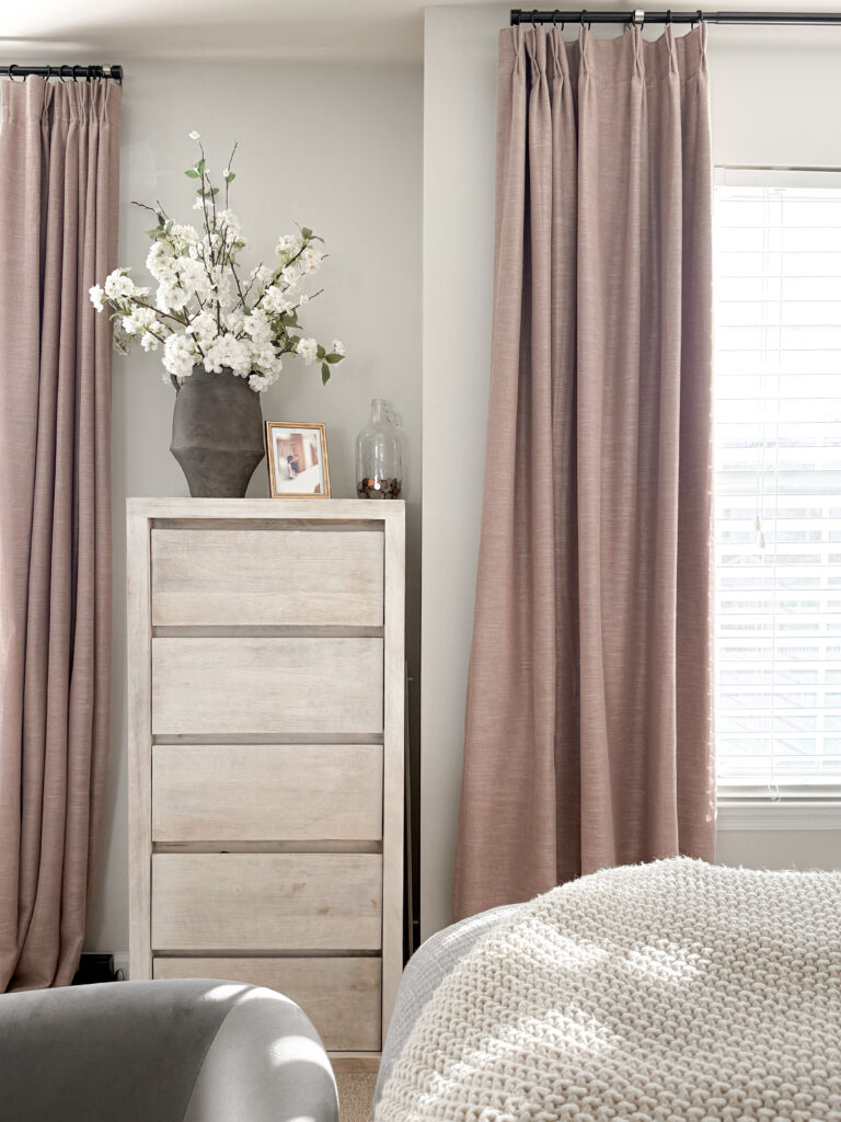 A soft, romantic corner of the bedroom featuring mauve curtains, a tall dresser, and a vase of fresh florals adding height and texture.