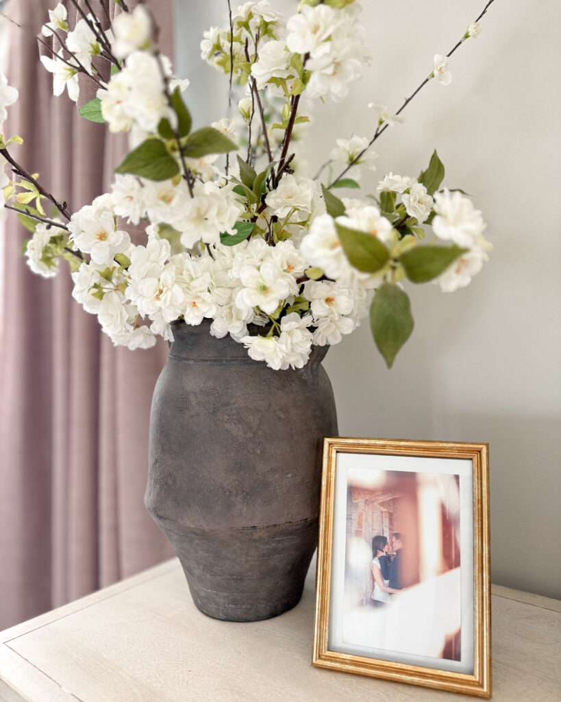 A dark stone vase filled with white blooms sits beside a gold picture frame, adding warmth and personality to the neutral decor.