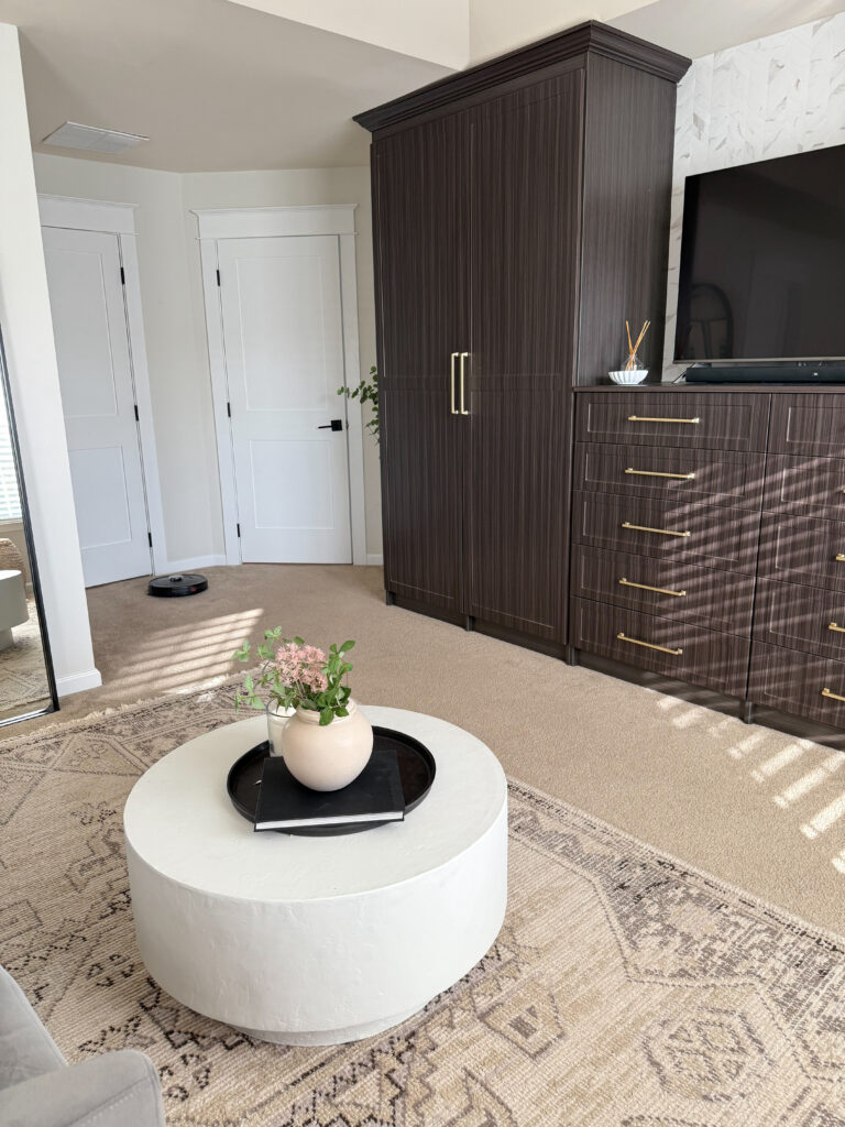 A close-up of the light round coffee table styled with a vase and tray, sitting on a neutral geometric rug beside the bed.