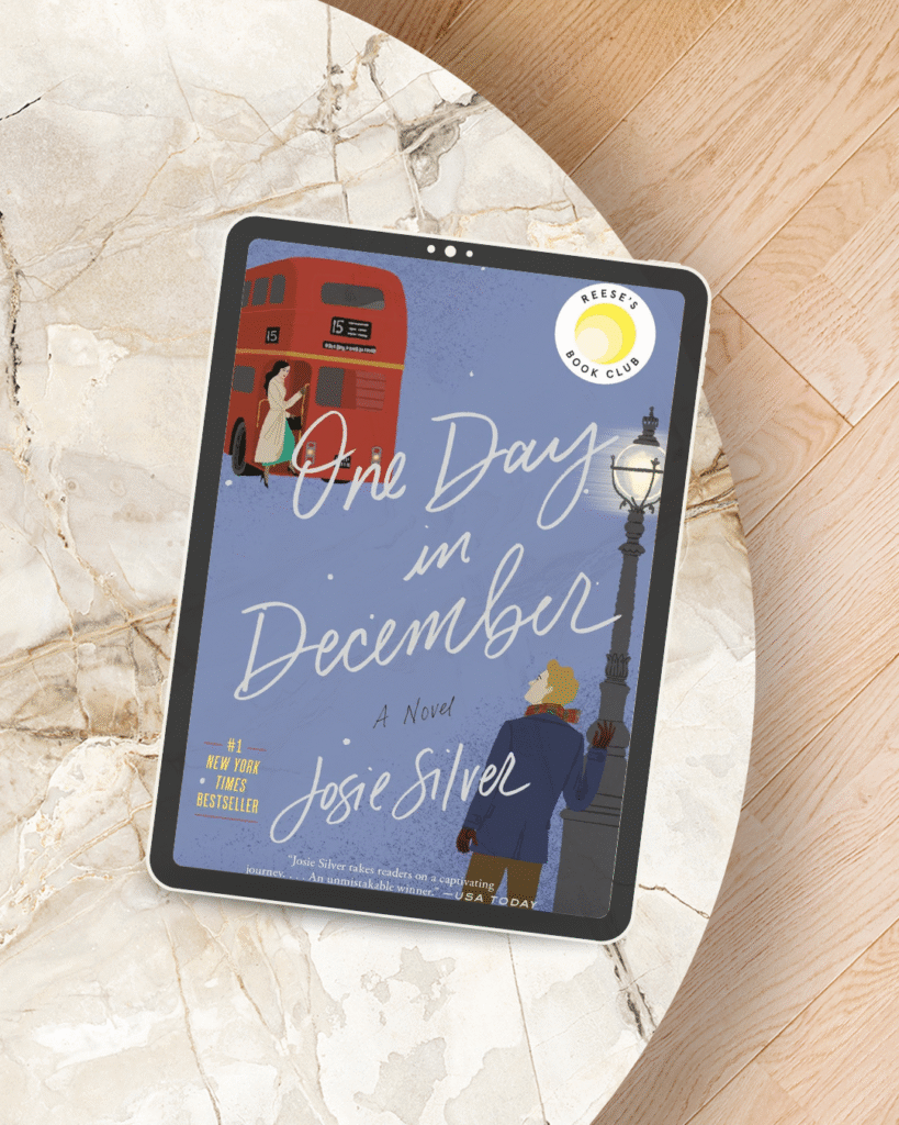 November's BookClub Digital tablet on a marble tabletop displaying the book cover of ‘One Day in December’ by Josie Silver, featuring an illustrated red London bus, a woman at the bus door, and a man standing under a streetlamp with Reese’s Book Club badge in the top corner.