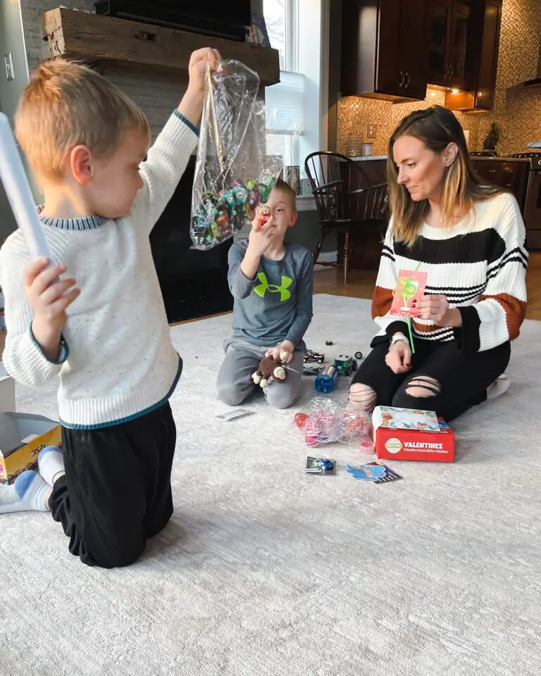 Two young boys sitting on a living room floor testing non-candy Valentine’s Day classroom favors while their mom looks on, with small toys, cards, and Valentine supplies spread out. Candy Free Valentine's