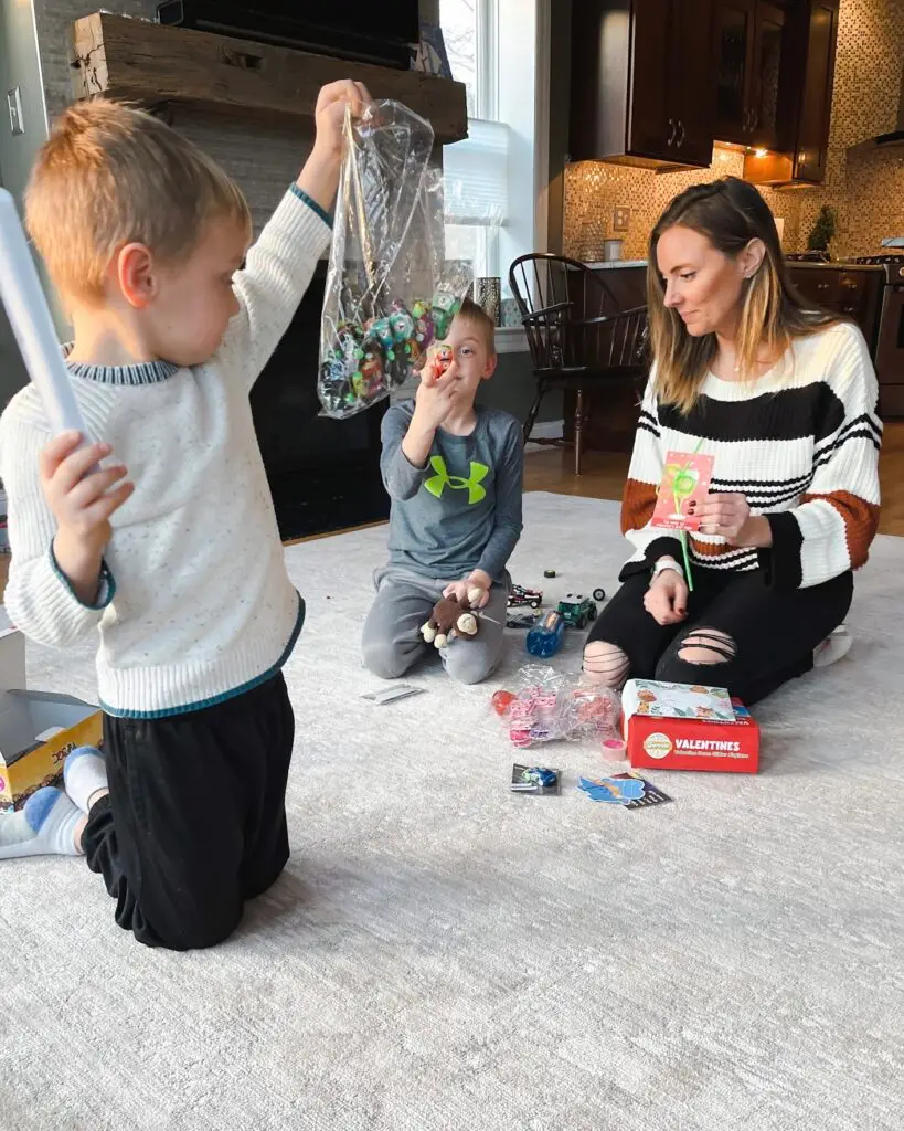 IMG_5108 1 - Sweet Motherly Two young boys sitting on a living room floor testing non-candy Valentine’s Day classroom favors while their mom looks on, with small toys, cards, and Valentine supplies spread out. Candy Free Valentine's