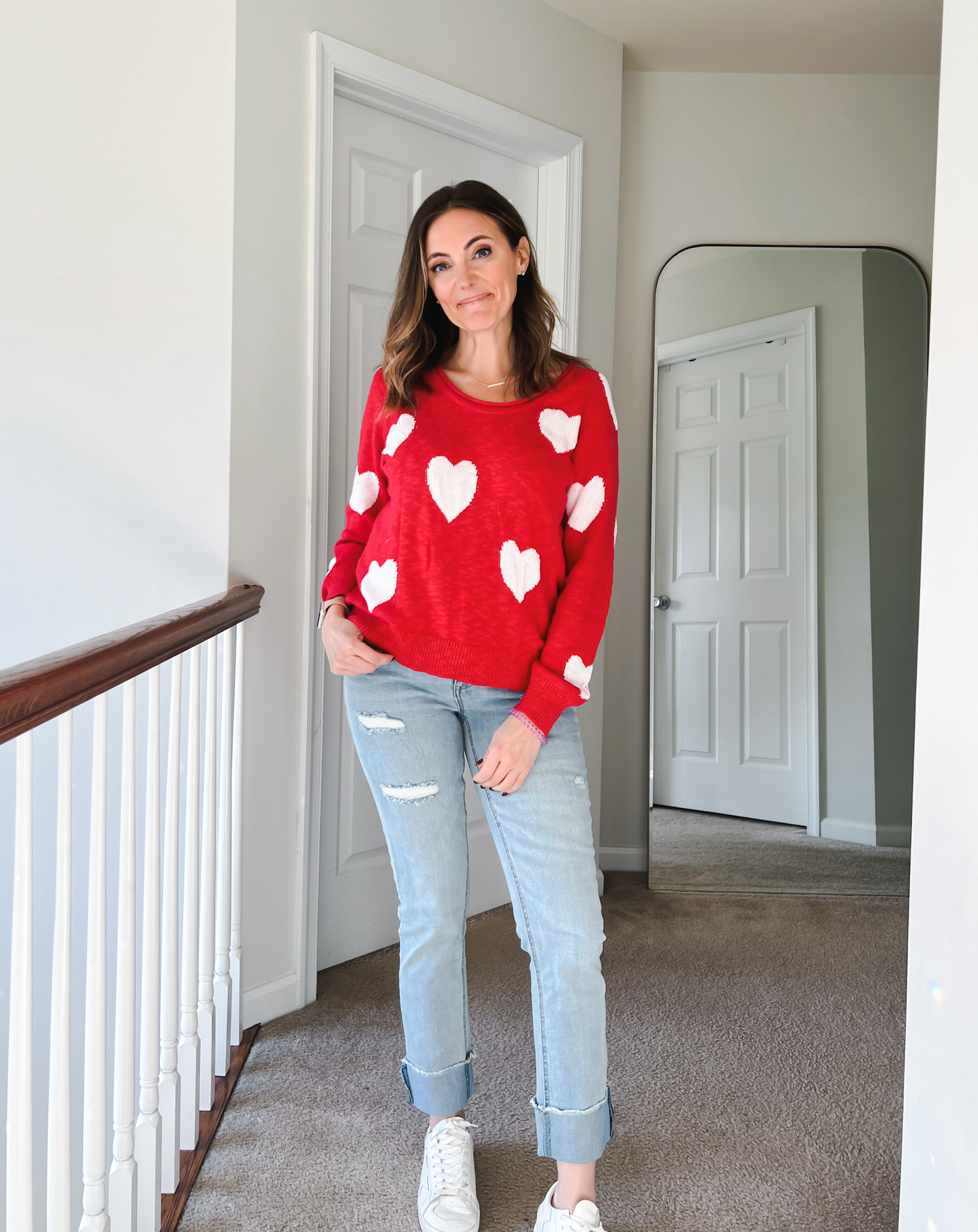 Woman wearing a red sweater with white heart print styled with cuffed light wash jeans and white sneakers for a casual Valentine’s Day look.