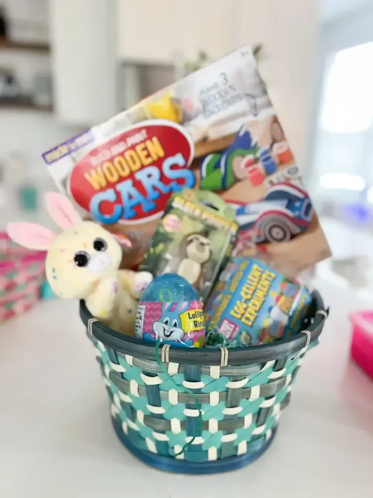 Blue and mint woven Easter basket for boys filled with wooden toy cars set, plush bunny, Egg-Cellent Experiments kit, lollipop candy egg, small animal toy, and assorted Easter candy and basket stuffers.