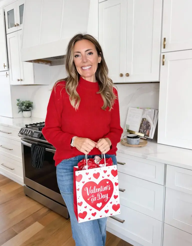 Woman wearing a red sweater and jeans holding a Valentine’s Day gift bag in a bright white kitchen, styled for a Valentine’s Day gift guide