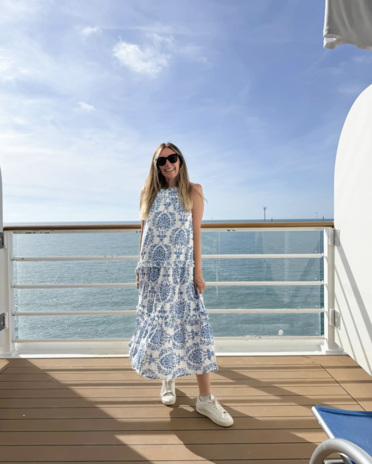 Woman wearing a blue and white printed tiered maxi dress with white sneakers and black sunglasses, standing on a balcony overlooking the ocean for a chic affordable resort wear look.