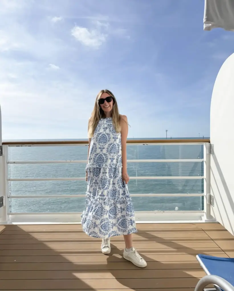 Screenshot 2026-02-02 at 100752 AM - Sweet Motherly Woman wearing a blue and white printed tiered maxi dress with white sneakers and black sunglasses, standing on a balcony overlooking the ocean for a chic affordable resort wear look.