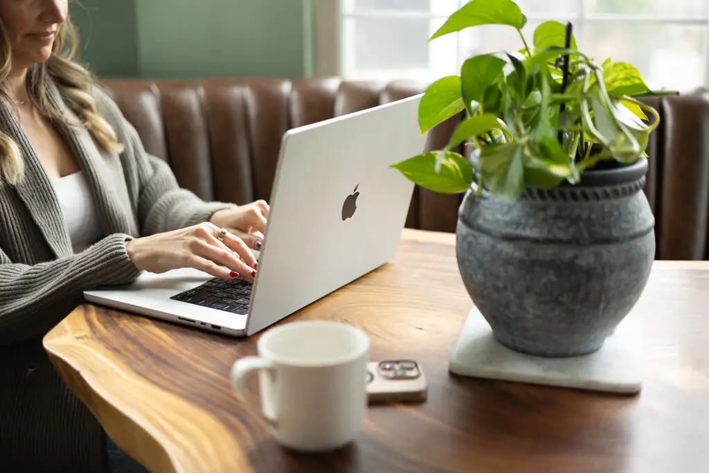 Woman working on a laptop at a wooden table with a potted plant and coffee mug, cozy home workspace