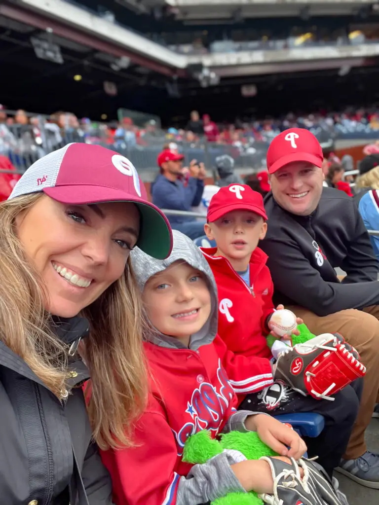Smiling woman and two children in red team caps at a baseball stadium, taking a selfie.