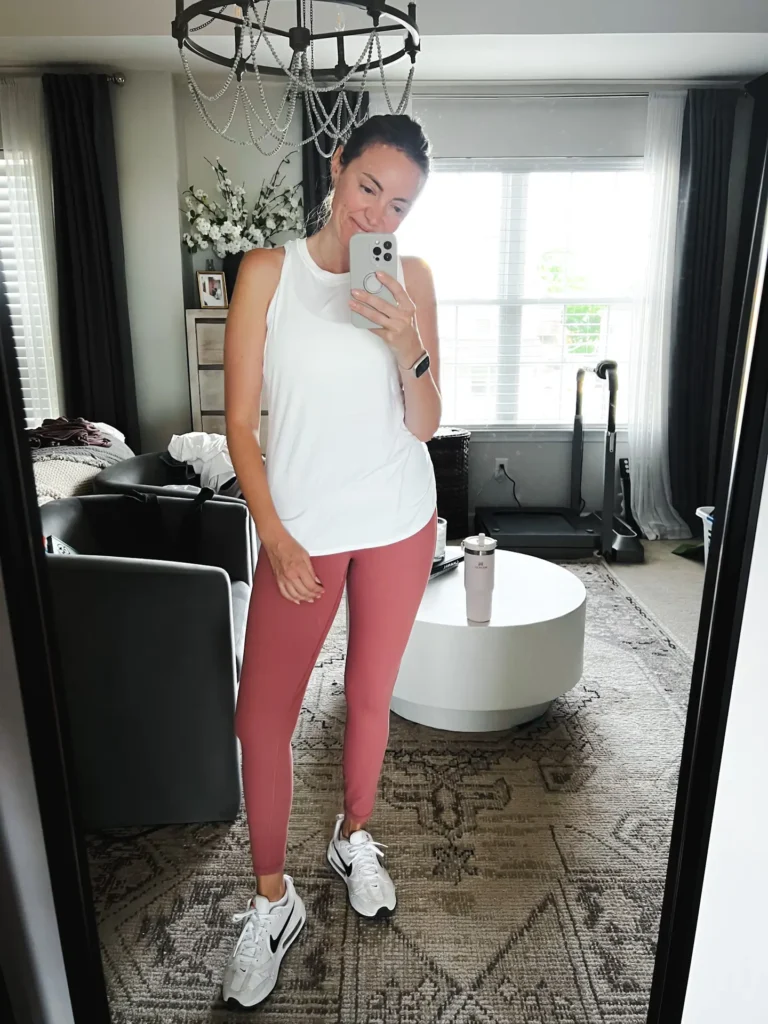 Woman taking a mirror selfie in a living room, wearing a white sleeveless top and pink leggings.