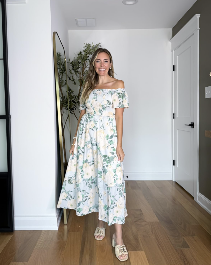 Smiling woman in a light blue floral off-shoulder dress standing in a bright room with a tall mirror and plant nearby.