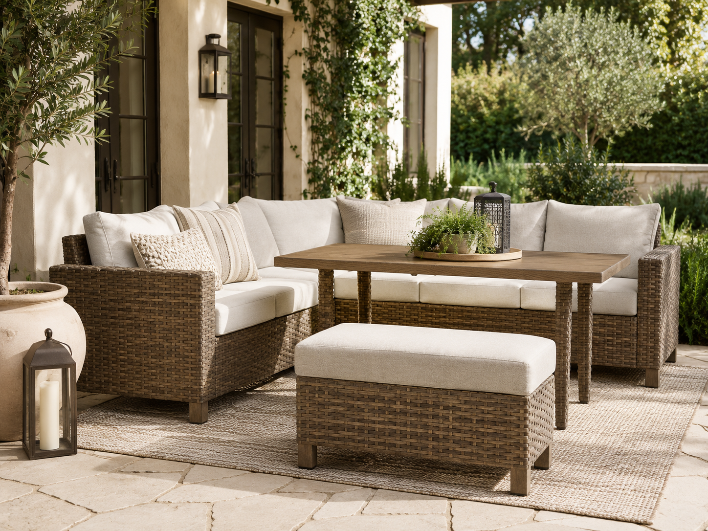 Outdoor wicker sectional sofa with beige cushions around a wooden coffee table on a stone patio, with lanterns and greenery nearby